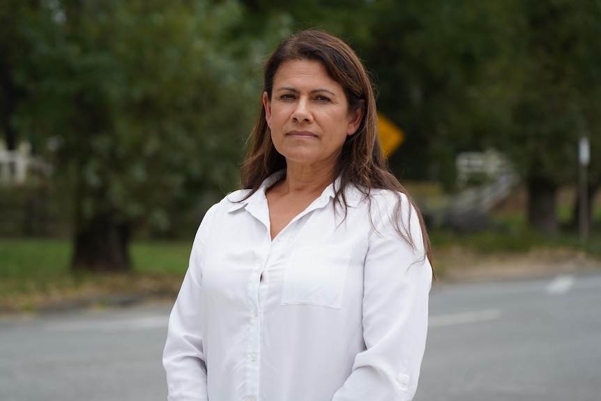 A woman wearing a white shirt standing at a slight angle, with a leafy street scene behind her.