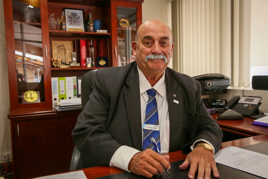 A smiling man with a grey moustache sits in his office.