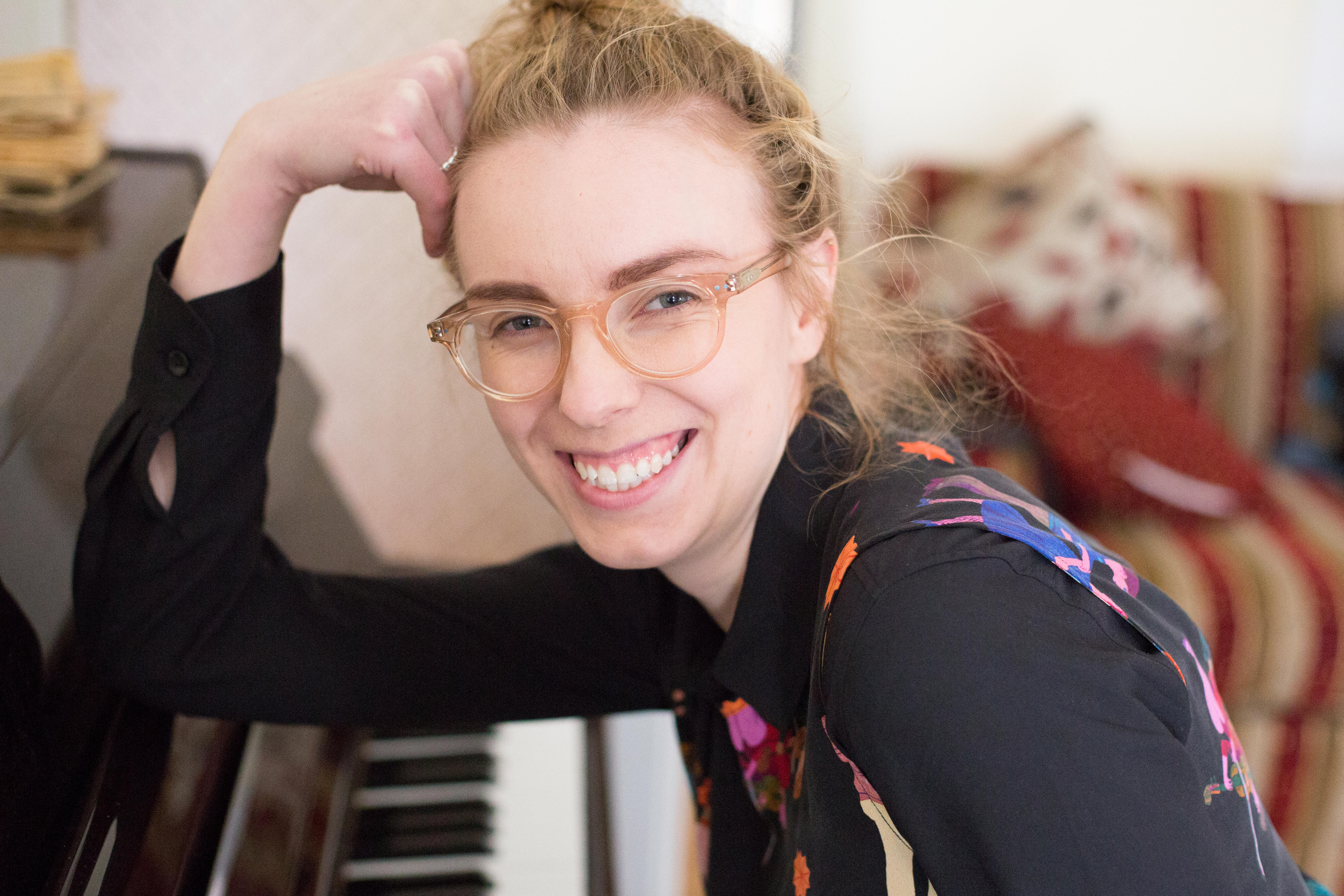 Zara Thompson leans her elbow against the open lid of an upright piano. She smiles at the camera and wears glasses.