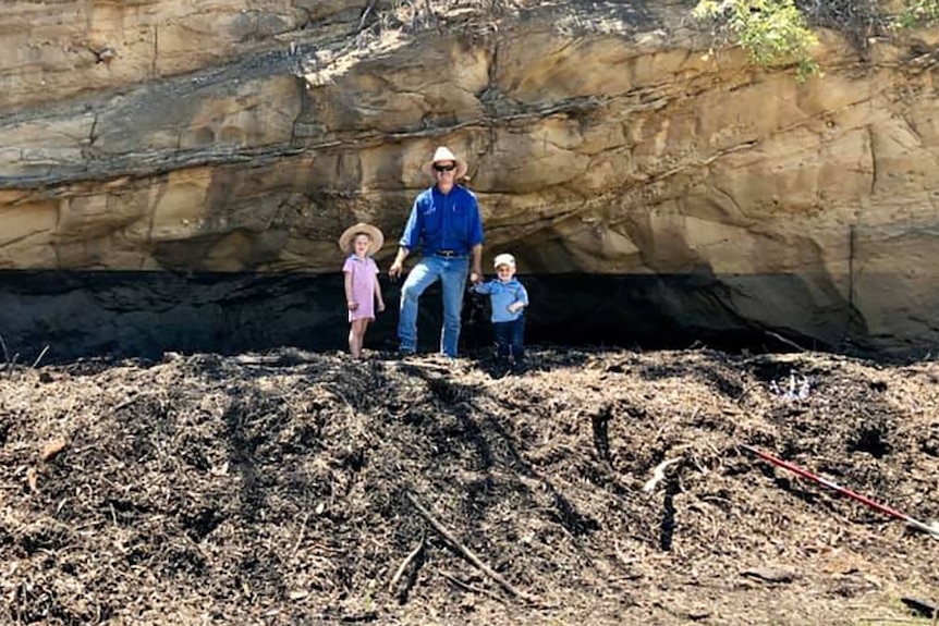 A man and two children standing on a mound of earth in front of a rock escarpment