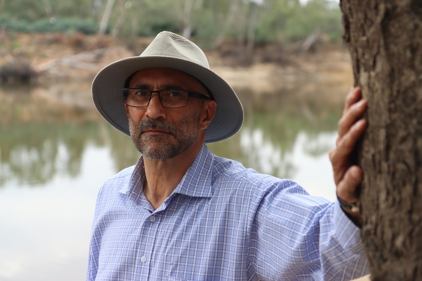 Ian Hamm wears a wide brim hat and blue shirt, he's leaning on a tree