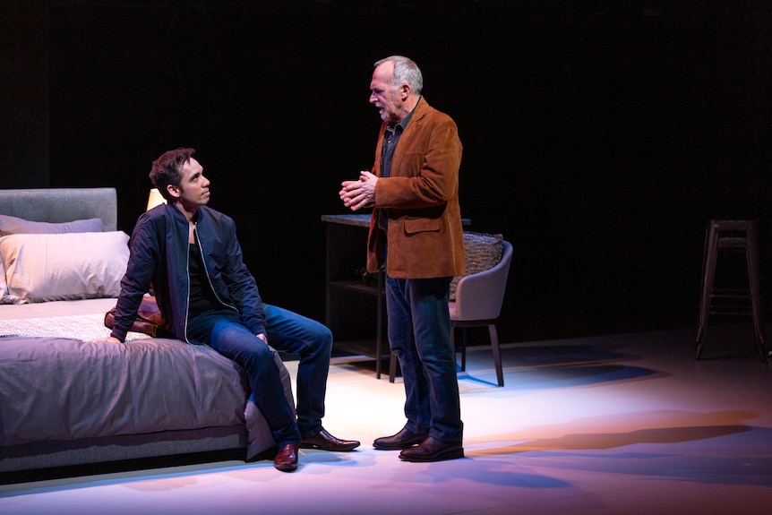 An actor on stage sits on a bed and listens as an older man, who is standing up, talks to him.