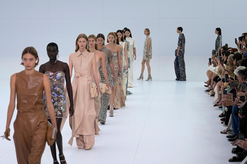 A line of models walk on an all white catwalk. 