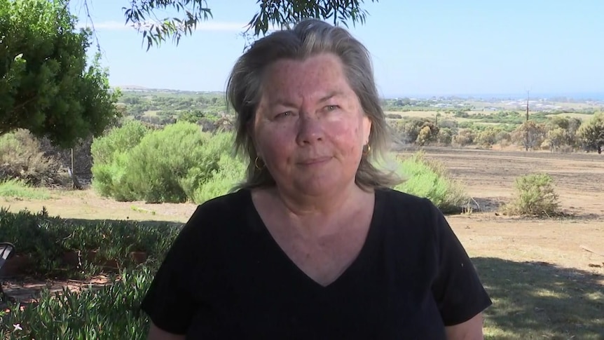 A woman stands outdoors with shrubbery and paddocks behind her