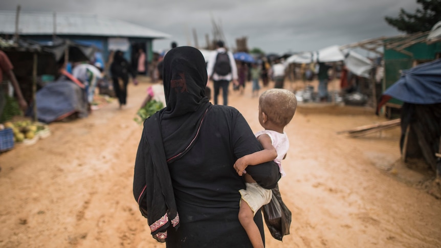 A woman in a hijab holds a baby and walks down a dusty road in a refugee camp