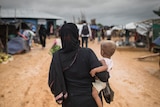 A woman in a hijab holds a baby and walks down a dusty road in a refugee camp