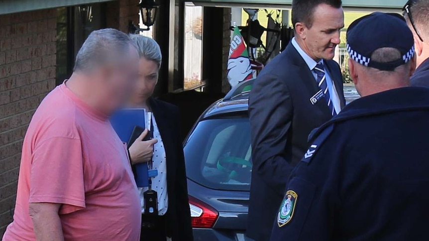 an older man in t-shirt and shorts with face pixelated is seen in the driveway of a home with police around him