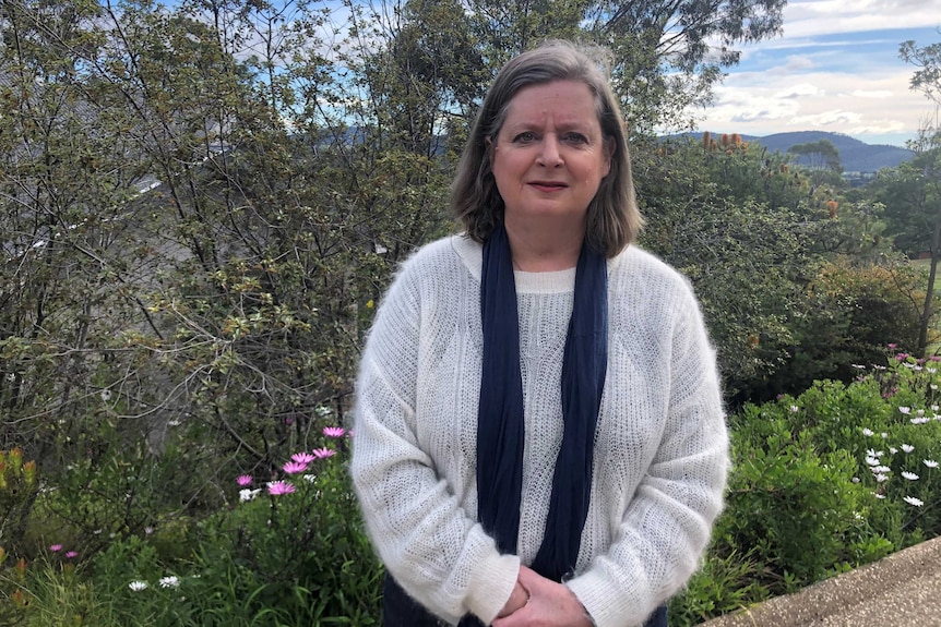 A woman wearing a white jumper stands in front of a garden.