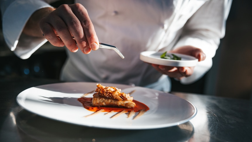 A chef's hands holding a pair of tweezers above a plate of food in a fine-dining restaurant
