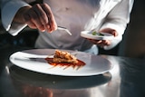 A chef's hands holding a pair of tweezers above a plate of food in a fine-dining restaurant