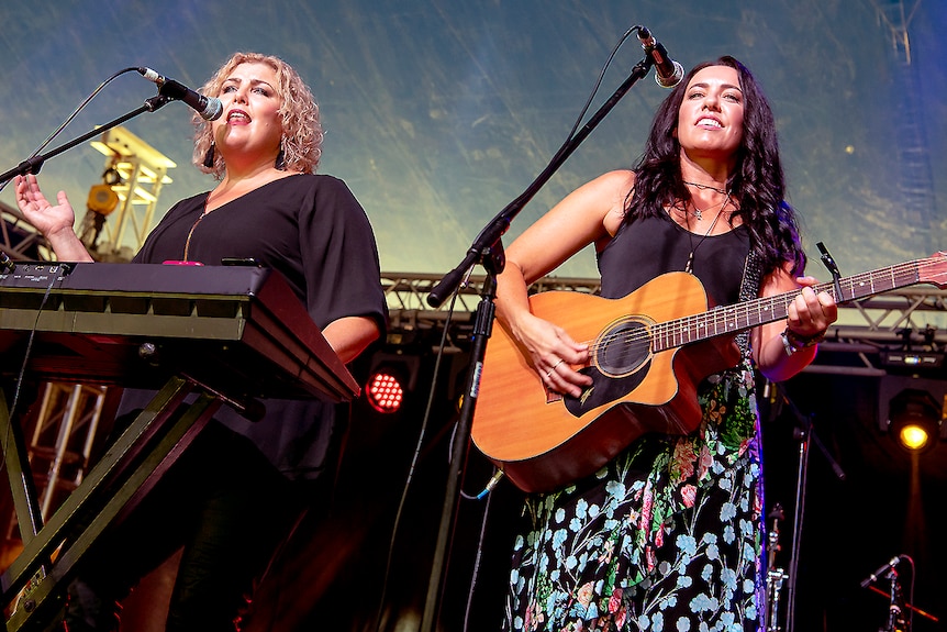 A woman with blonde curly hair sings in front of a keyboard and a woman with long dark hair plays guitar and sings.