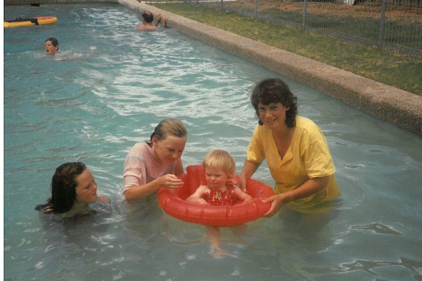 A small child sits in a flotation ring in the swimming pool and is supported by two girls and a woman at Tirlta Station.