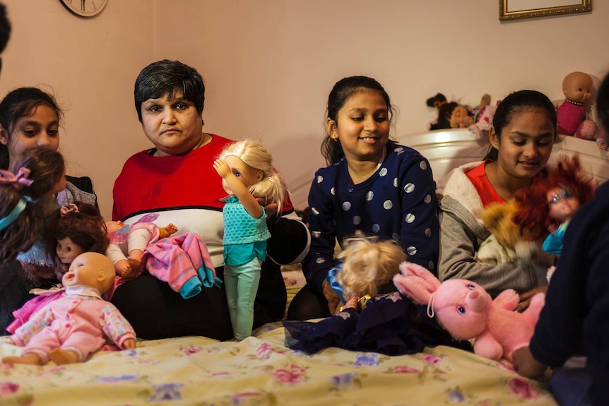 A middle-aged Indian woman sits on a bed with three teenaged relatives, as they play with dolls.
