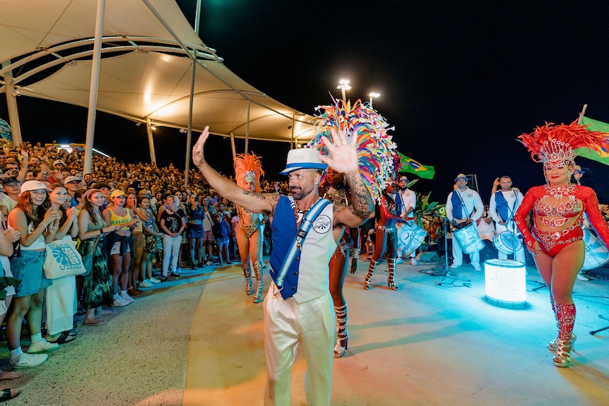 man waves to a large crowd with samba dancers and drummers behind him 