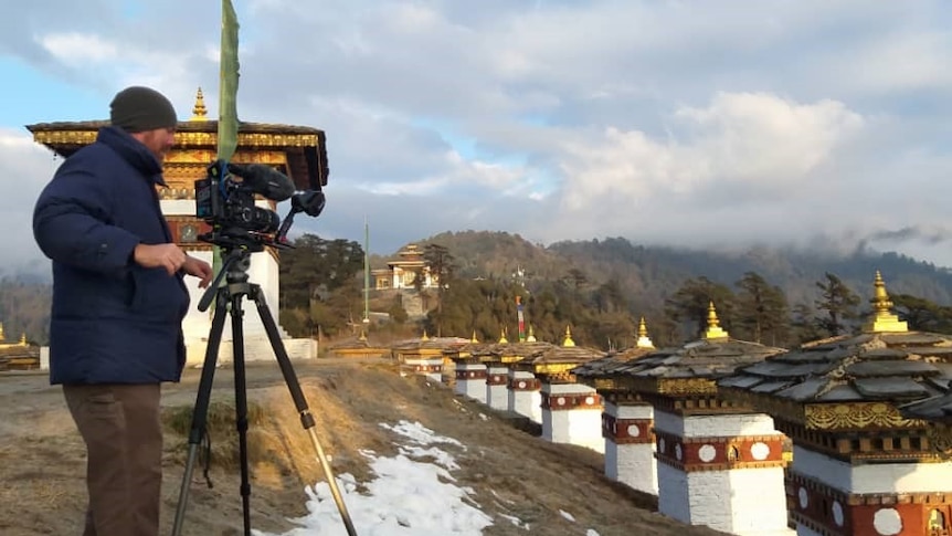 Smith standing with camera on tripod pointing towards mountains with low cloud and traditional buildings in foreground.