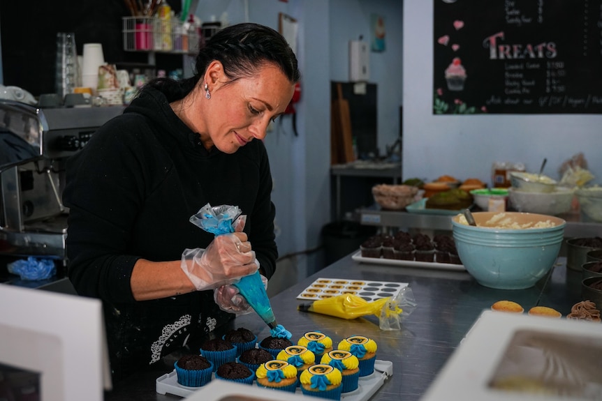 A woman piping blue and yellow icing on cupcakes.
