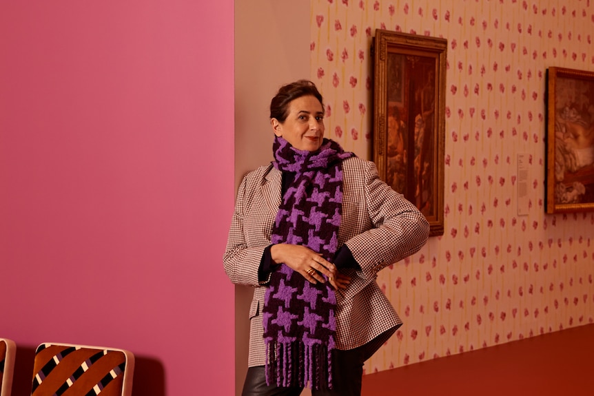 A woman with dark hair stands smiling in a gallery space with vividly coloured walls, with a painting in the background.