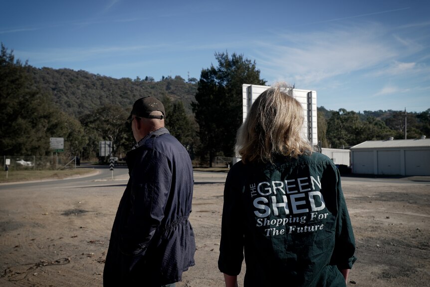 A woman wearing a shirt that says 'The Green Shed' and a man in a cap stand on dirt next to a road.
