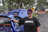 A woman with short purple hair smiles,  wears dark sunglasses and a black t-shirt, in front of a purple car.