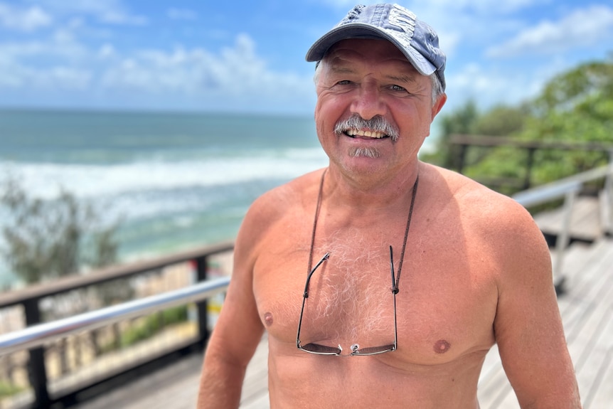 a man smiles at the camera standing shirtless on a beach viewing platform. he has glasses around his neck and a cap on