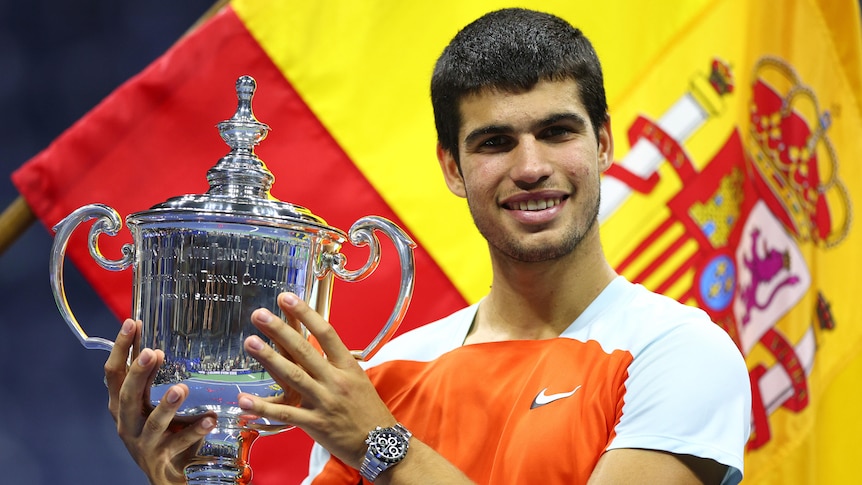 Spain's Carlos Alcaraz smiles as he holds up the trophy at the US Open, with a Spanish flag behind him.