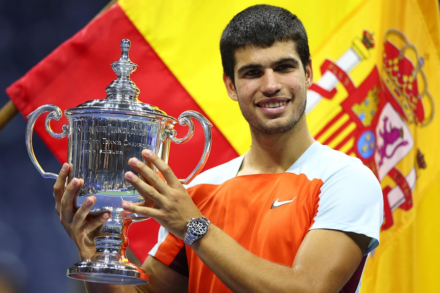 El español Carlos Alcaraz sonríe mientras sostiene el trofeo en el US Open, con la bandera española detrás de él.