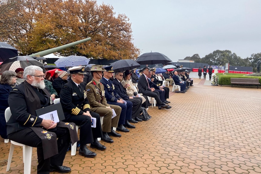 A group of people, some in military uniforms, sit in the rain at a service.