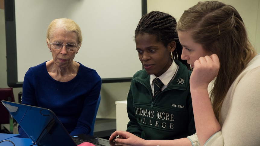 A teacher sits with two students who are learning computer skills.