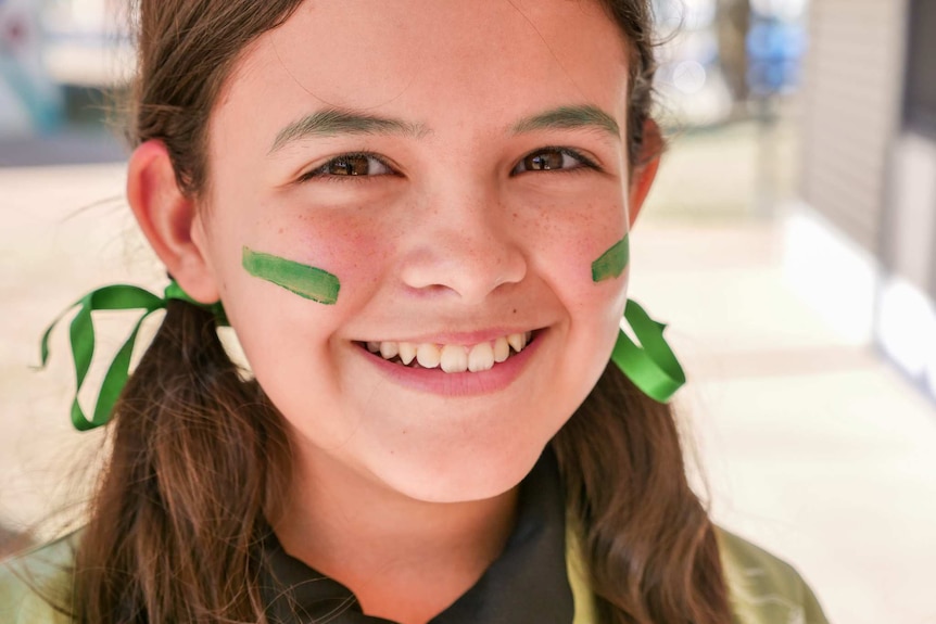 A smiling young girl wearing green face paint looks at the camera. She has brown eyes and hair and freckles.