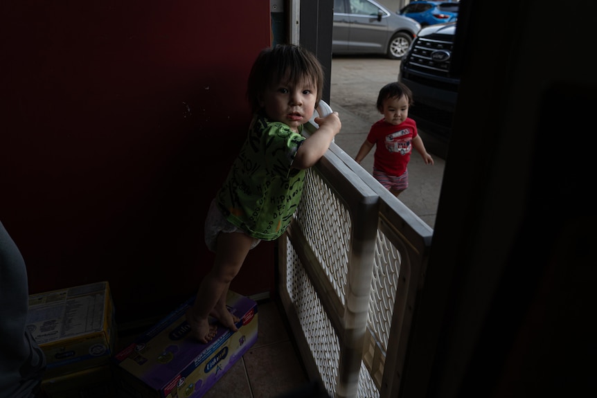 A toddler in a green shirt leans out over a baby gate at the door of a motel room. A toddler in a red shirt runs past the room.