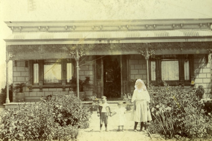 Faded black and white photo of a federation house with a woman and child standing in its front yard, with bushes on each side.