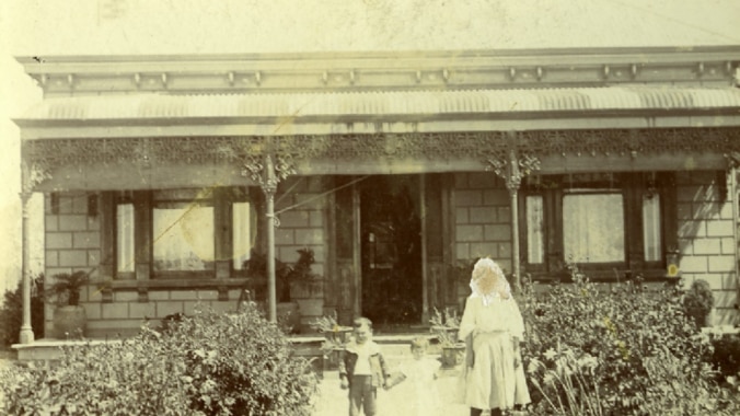 Faded black and white photo of a federation house with a woman and child standing in its front yard, with bushes on each side.