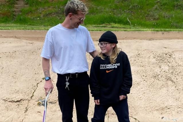 Jayden stands next to his son on the skateboard, holding his shoulder.