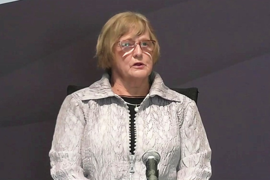 An older woman sits in a witness chair and speaks in front of a microphone.