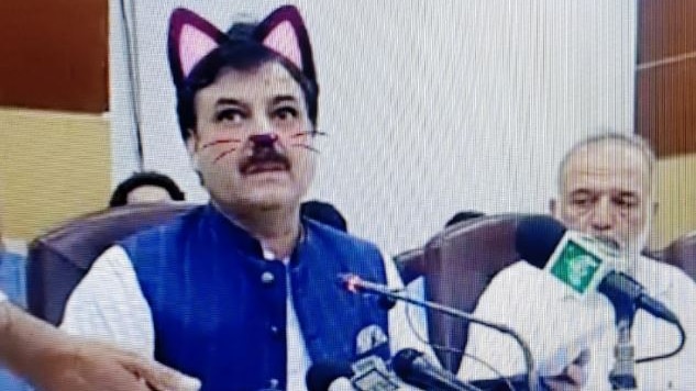 A man sitting at a desk several microphones pointed at him. A pair of cat ears, nose and whiskers appear over his face.