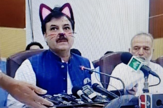 A man sitting at a desk several microphones pointed at him. A pair of cat ears, nose and whiskers appear over his face.