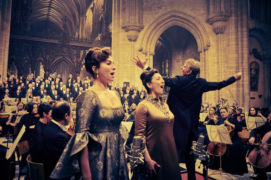 A musical performance in a cathedral with a choir, orchestra, conductor and two female singers in the foreground