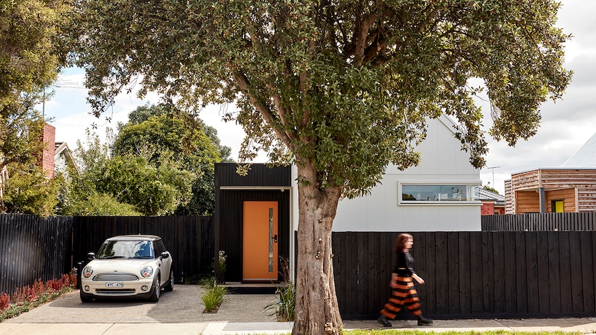 A street view of a small white and black home with an orange door behind a black fence. There's a Mini Cooper in the driveway.