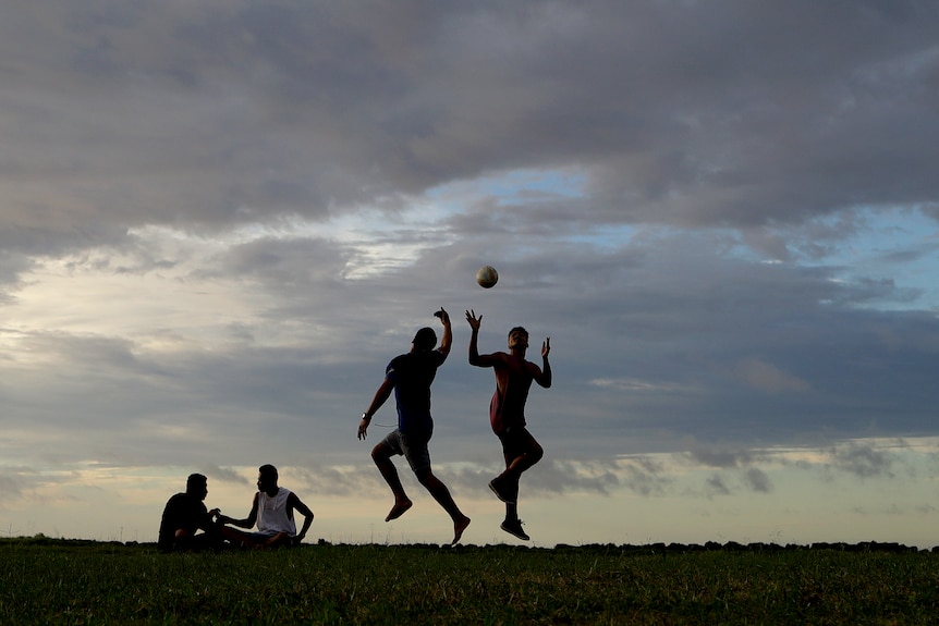 A silhouette of young men playing soccer jumping with clouds in the background.