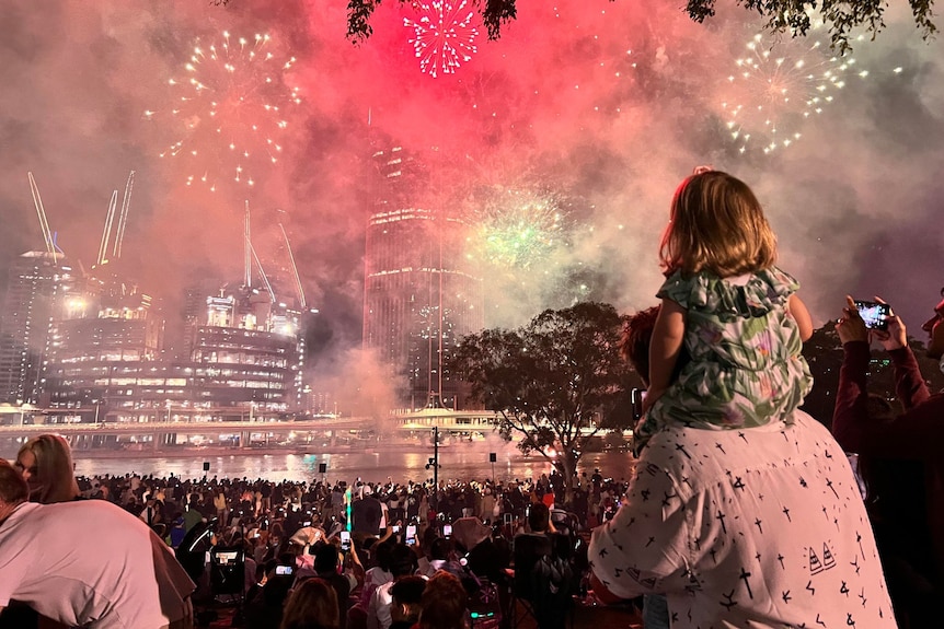 A young girl sits on a man's shoulders watching fireworks.