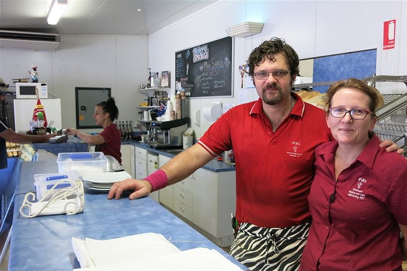 Bakers John and Estelle Carter stand at the counter of a bakery with serious expressions. Behind them, a customer is served.