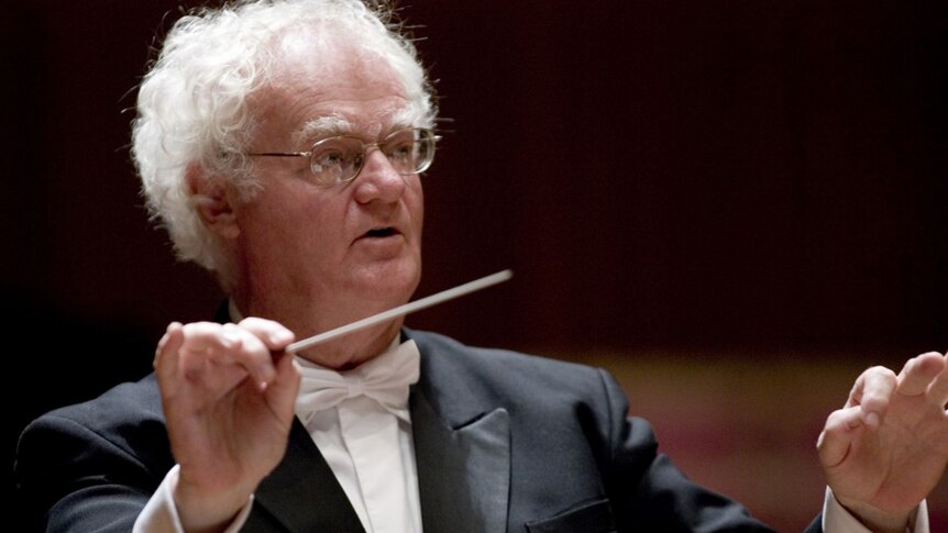 A photo of Richard Gill in a tuxedo with white tie holding a conductor's baton.