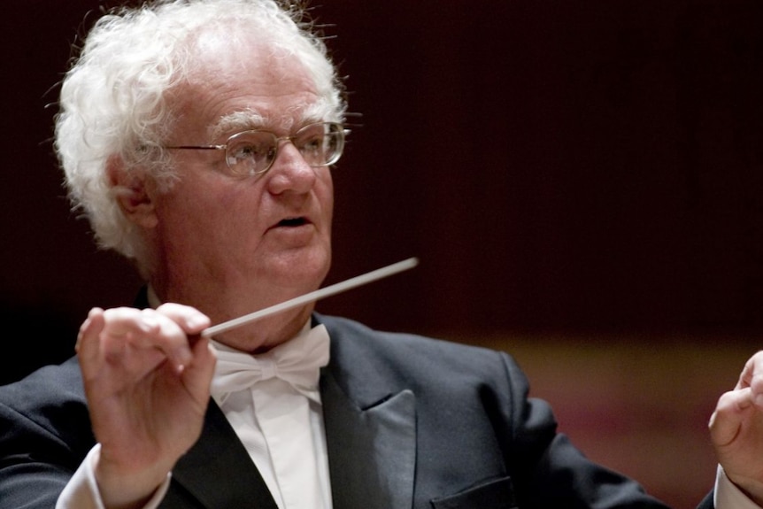 A photo of Richard Gill in a tuxedo with white tie holding a conductor's baton.