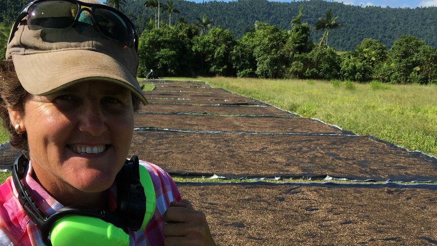 A young farmer leans on her rake in front of large black mats full of black peppercorns drying in the hot sun