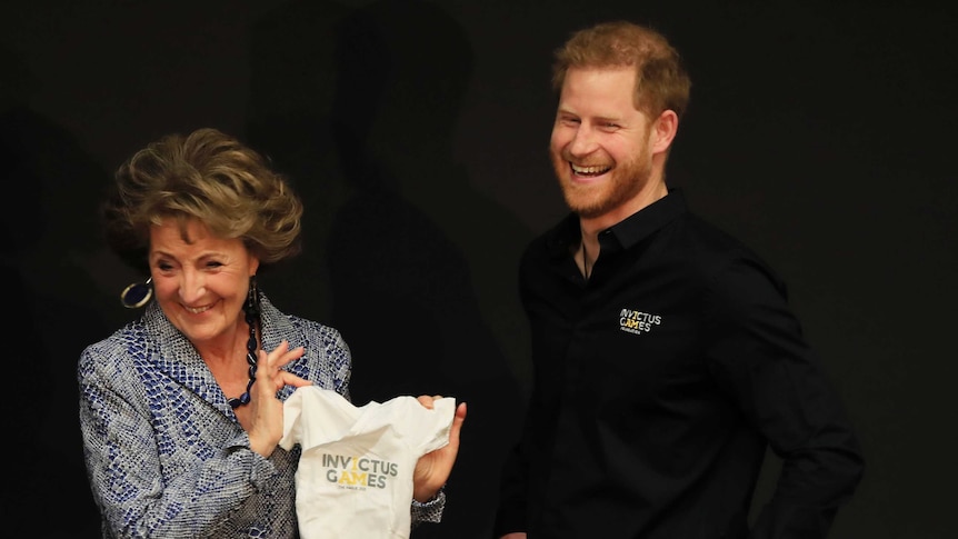 Against a black background, Prince Harry laughs as Princess Margriet holds a white baby onesie.