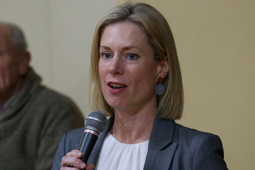 Rebecca White, wearing a grey blazer and white top, holds a microphone