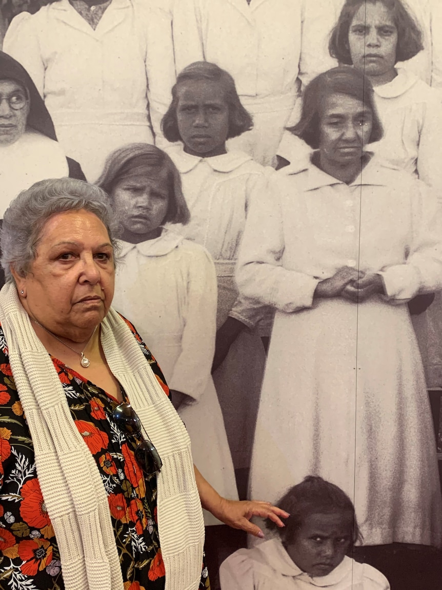 Beverley Port-Louis standing in front of large black and white photograph of girls and nuns at St Joseph's orphanage