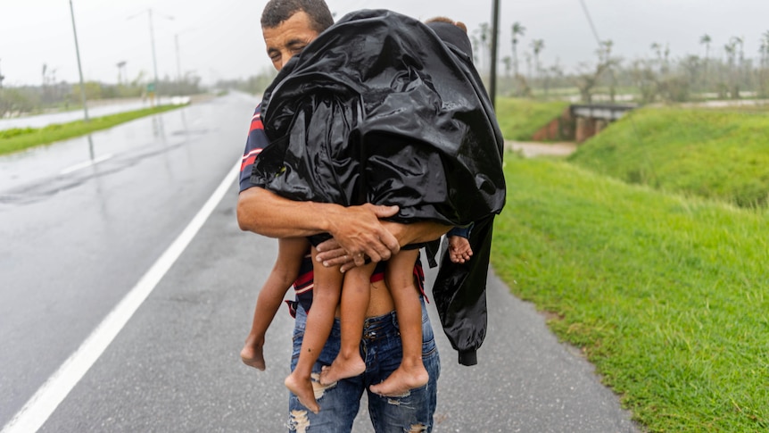 man carries small children whose feet are visible under a plastic black cover, lashed with rain