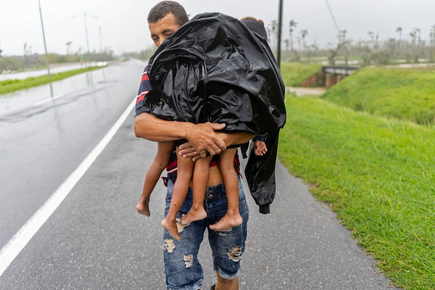 man carries small children whose feet are visible under a plastic black cover, lashed with rain