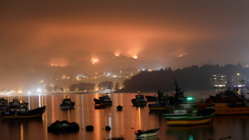 A harbour with several boats in it with a red smokey haze over it with flames in the background